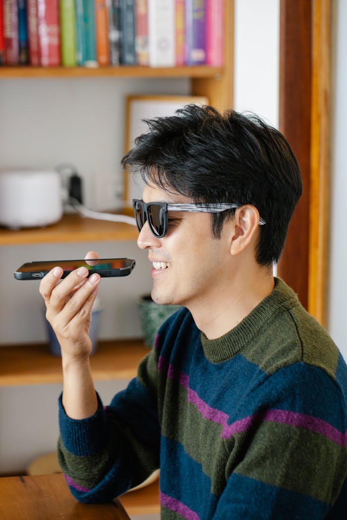our-experience A smiling man wearing sunglasses uses voice command on a smartphone indoors.
